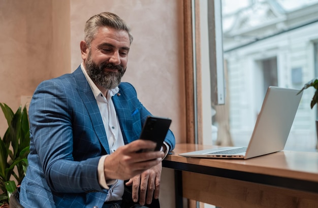 Un felice uomo d'affari maturo che utilizza il suo telefono mentre lavora su un computer portatile in un ristorante caffetteria