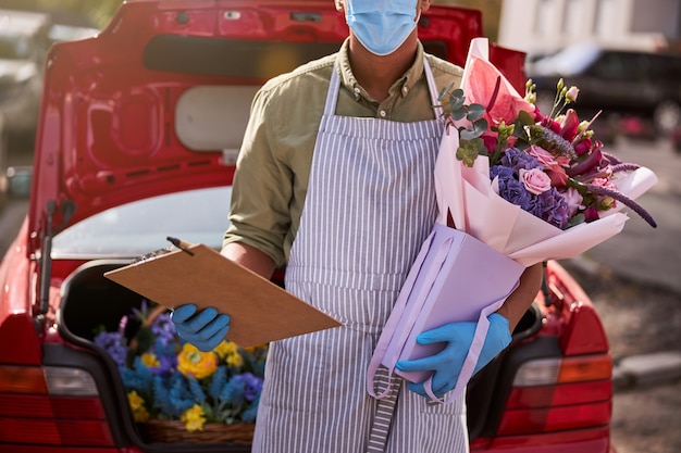 un fiorista multietnico che tiene un blocco per appunti con una penna e un bouquet confezionato accanto a una macchina