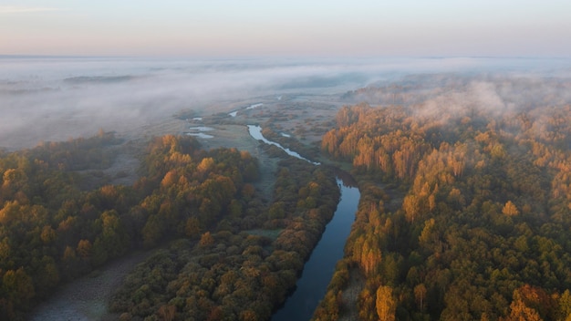 Un fiume tra campi e foreste tra nuvole e nebbia con le prime gelate durante la mattina d'autunno d'oro, vista dall'alto da un drone