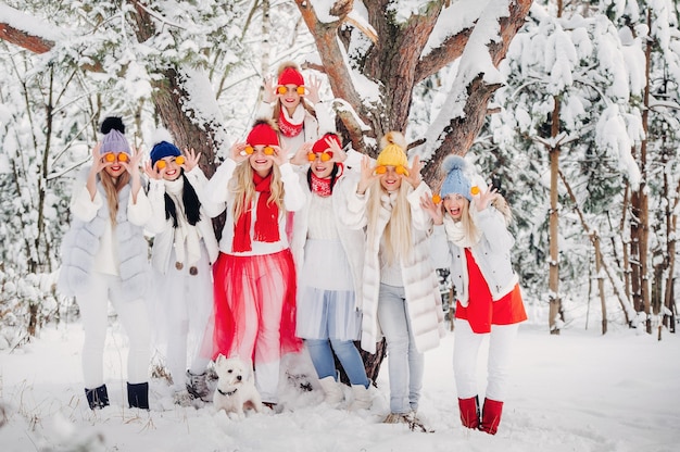 Un folto gruppo di ragazze con i mandarini sono in piedi nella foresta invernale. Ragazze in abiti rossi e bianchi con frutta in una foresta innevata.