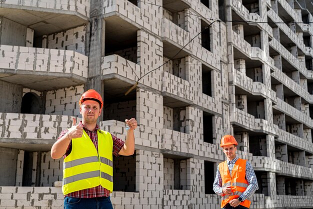 Un forte lavoratore in uniforme e casco tiene le chiavi del nuovo edificio e un altro lavoratore in uniforme e casco si trova più lontano da lui nel cantiere. Concetto di costruzione