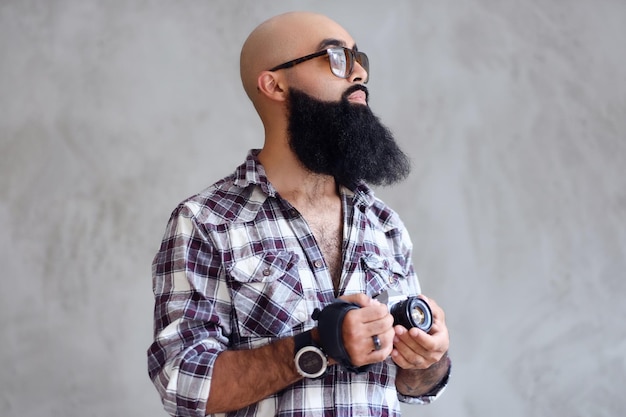 Un fotografo amatoriale hipster barbuto con tatuaggi sulle braccia, vestito con una maglietta in pile tiene una fotocamera DSLR compatta su sfondo grigio.