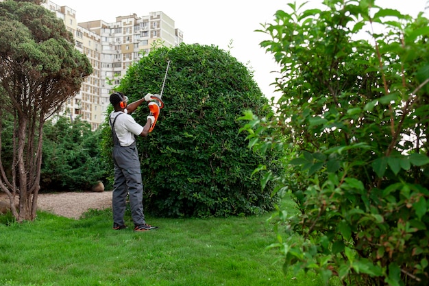 Un giardiniere in uniforme taglia cespugli. Un uomo afroamericano con occhiali e cuffie lavora in giardino.