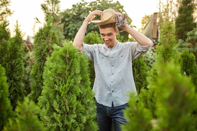 Un giardiniere sorridente con un cappello di paglia si trova nel vivaio con molti thuja in una calda giornata di sole.