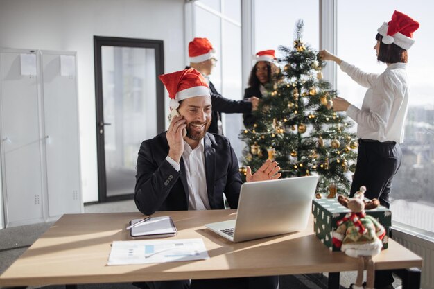 Un gioioso uomo d'affari caucasico che parla su uno smartphone mentre mostra un gesto di saluto usando un computer portatile sullo sfondo dei colleghi che decorano l'albero di Natale