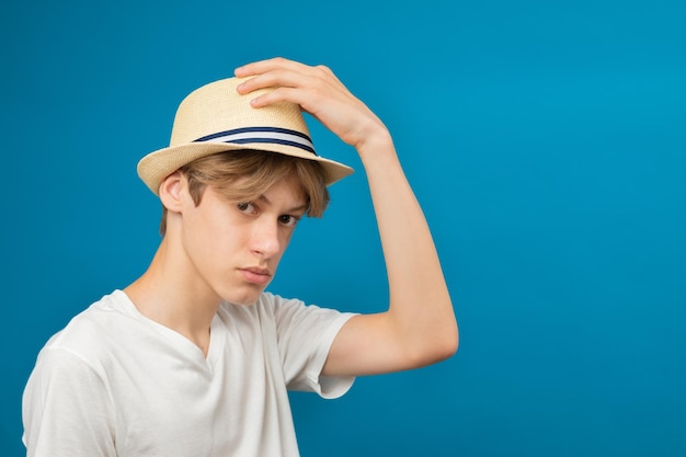 Un giovane adolescente che indossa una maglietta bianca e tiene in mano un cappello bianco in uno studio su sfondo blu Ritratto a mezzo busto di un giovane ragazzo in studio