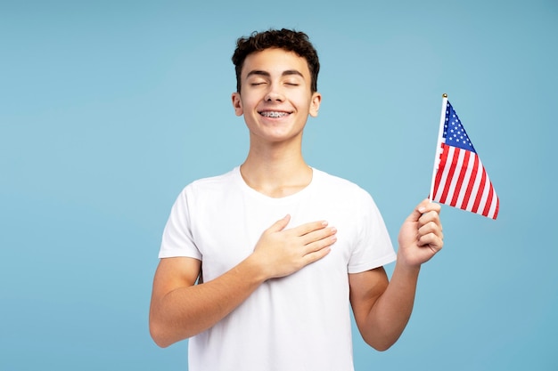 Un giovane adolescente patriota sorridente che tiene la bandiera americana con gli occhi chiusi e la mano sul cuore.