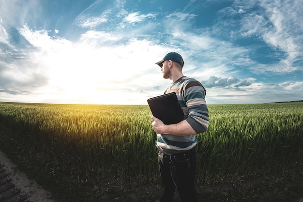 Un giovane agronomo tiene in mano una cartella su un campo di grano verde. Un contadino prende appunti sullo sfondo di terreni agricoli durante il tramonto. Uomo con un berretto con una cartella di documenti