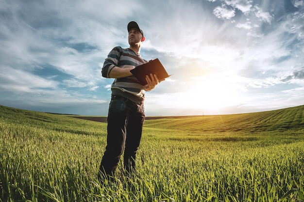 Un giovane agronomo tiene in mano una cartella su un campo di grano verde. Un contadino prende appunti sullo sfondo di terreni agricoli durante il tramonto. Uomo con un berretto con una cartella di documenti