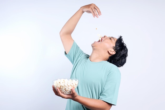 Un giovane asiatico divertente che mangia e si diverte a mangiare popcorn su uno sfondo bianco.