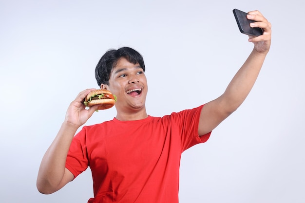 Un giovane asiatico felice tiene in mano un grande hamburger e si fa un selfie con uno smartphone