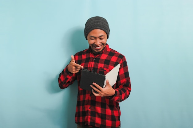 Un giovane asiatico sorridente con un berretto e una camicia di flanella scozzese rossa sta indicando un libro in mano mentre si trova su uno sfondo blu