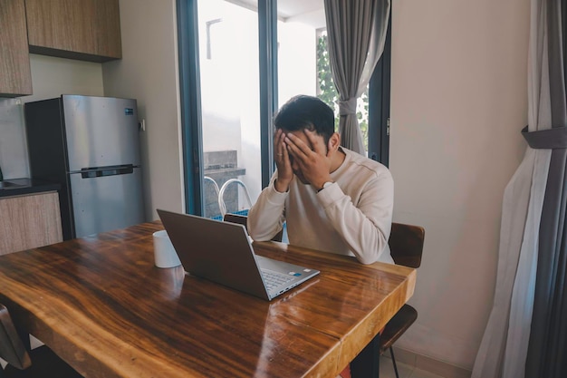 Un giovane asiatico trascorre il suo tempo a casa seduto nella sala da pranzo a lavorare sul suo laptop sentendosi frustrato