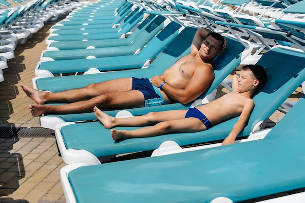 Un giovane atletico e suo figlio sorridono felici e prendono il sole su un lettino in una giornata di sole in hotel Buone vacanze in famiglia in un hotel del resort Vacanze estive e turismo