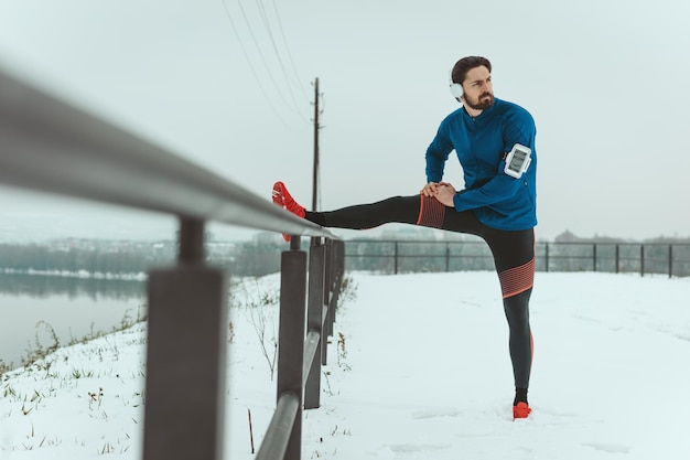 Un giovane attivo, con le cuffie sulle orecchie, si sta allungando e facendo esercizi in un luogo pubblico durante l'allenamento invernale all'aperto.