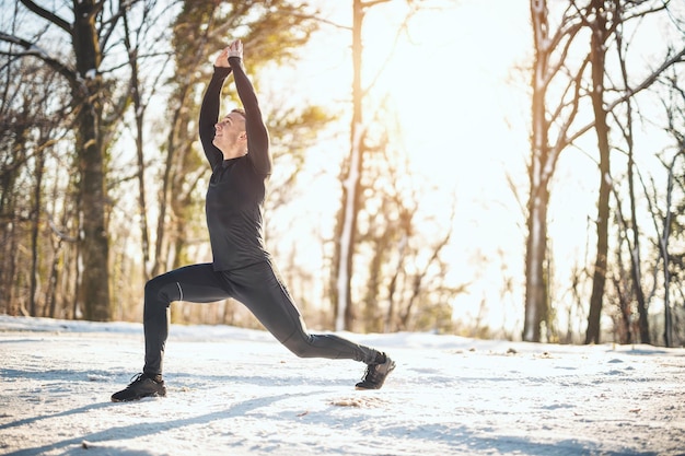 Un giovane attivo sta facendo esercizi di stretching nella strada forestale durante l'allenamento invernale all'aperto.