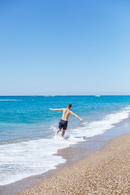 Un giovane attraente con un bel corpo in pantaloncini corti cammina lungo la spiaggia sabbiosa e si gode le vacanze estive