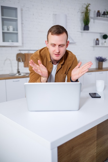 Un giovane attraente in abiti casual seduto in cucina utilizzando un computer portatile. Lavoro da casa, flusso di lavoro remoto.