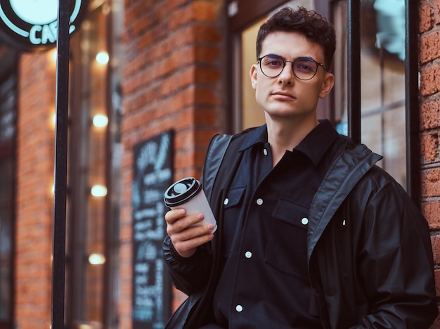 Un giovane bell'uomo che indossa una giacca con in mano un caffè da asporto mentre è in piedi vicino a un bar fuori.