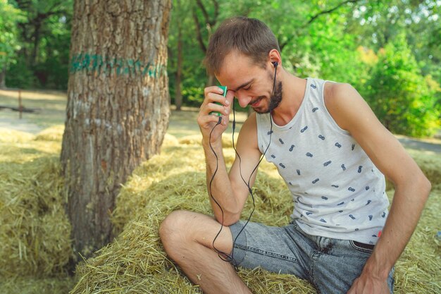 Un giovane bell'uomo con la barba in una maglietta è seduto nel fieno, ascolta musica e canta nel parco