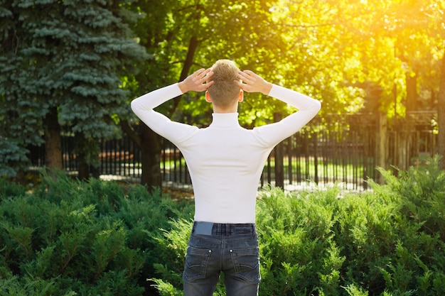Un giovane biondo, vestito con un maglione bianco e jeans neri, sta con le spalle alla telecamera, tenendo la testa tra le mani sullo sfondo di un parco cittadino. Concetto di foto.