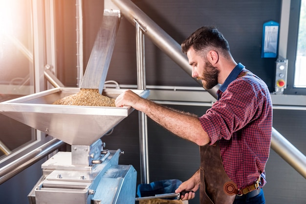 Un giovane birraio in un grembiule in pelle controlla la macinazione dei semi di malto in un mulino in una moderna fabbrica di birra