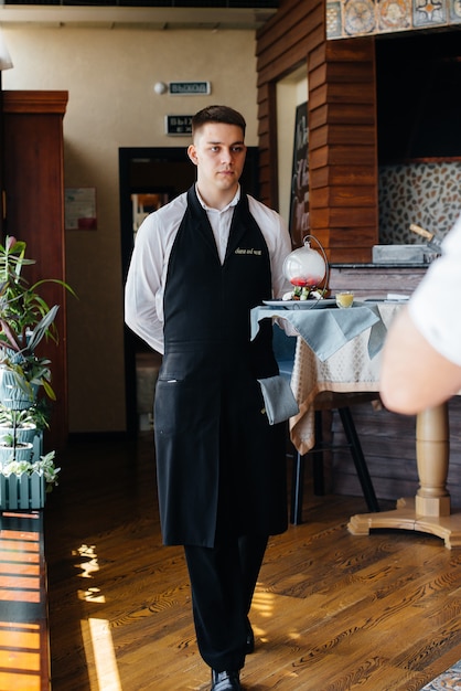 Un giovane cameriere con un'elegante uniforme in piedi con un piatto squisito su un vassoio vicino al tavolo in un bellissimo ristorante ravvicinato. Attività di ristorazione, di altissimo livello.