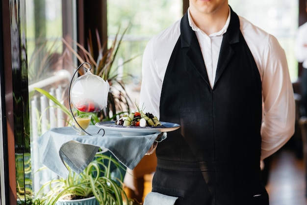 Un giovane cameriere con un'elegante uniforme in piedi con un piatto squisito su un vassoio vicino al tavolo in un bellissimo ristorante ravvicinato. Attività di ristorazione, di altissimo livello.