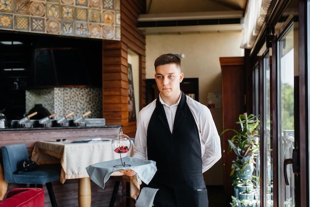 Un giovane cameriere con un'elegante uniforme in piedi con un piatto squisito su un vassoio vicino al tavolo in un bellissimo ristorante ravvicinato. Attività di ristorazione, di altissimo livello.