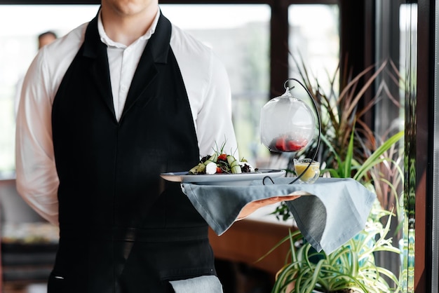 Un giovane cameriere in un'elegante uniforme sta con un piatto squisito su un vassoio vicino al tavolo in un bellissimo ristorante in primo piano Attività di ristorazione di altissimo livello