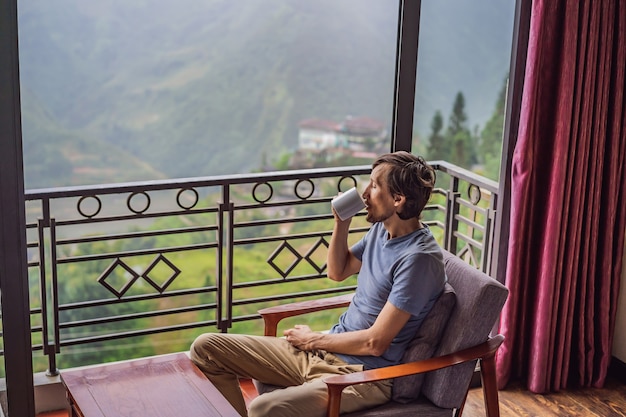 Un giovane che tiene in mano una tazza di caffè mentre è seduto su una sedia sul balcone guardando le montagne e il verde