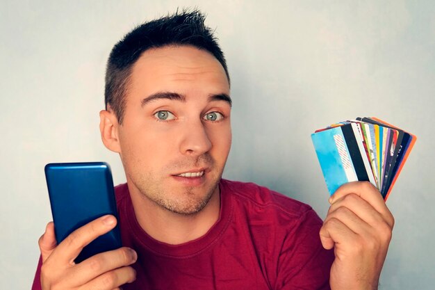 Un giovane che tiene uno smartphone in una mano nell'altra molte diverse carte di debito su sfondo blu scegliendo la carta bancaria per il pagamento nel negozio online