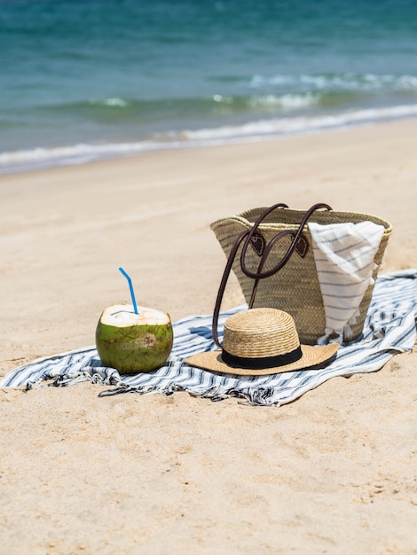 Un giovane cocco fresco è pronto da mangiare e una borsa di paglia e un cappello di paglia da donna su un asciugamano su una spiaggia sabbiosa contro un mare blu. Concetto di viaggio vacanza tropicale. Copia spazio