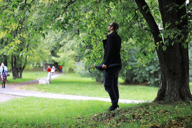 Un giovane con gli occhiali cammina nel parco con un ombrello durante la pioggia.