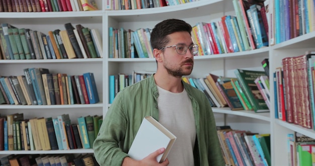 Un giovane con gli occhiali e una camicia verde con un libro in mano cammina vicino agli scaffali con un libro in biblioteca Concetto di biblioteca universitaria