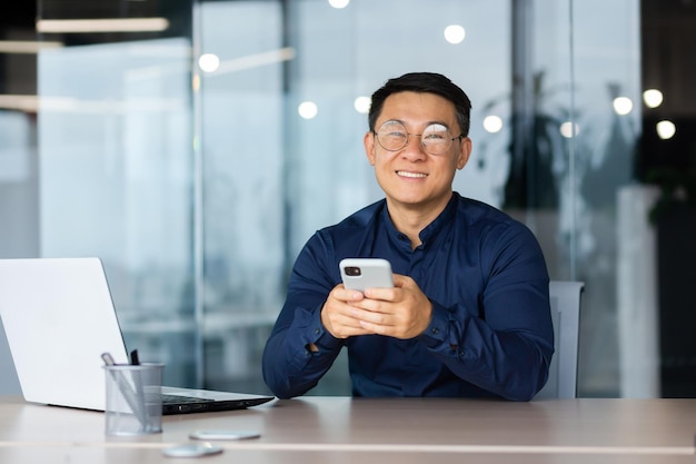 Un giovane con gli occhiali siede in ufficio a un tavolo con un laptop che tiene in mano un telefono sorridendo