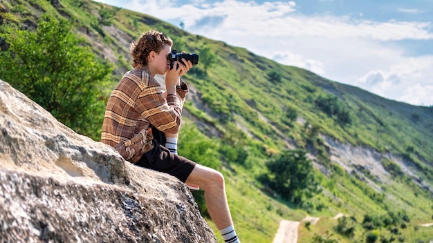 Un giovane con i capelli ricci che scatta foto usando la macchina fotografica nella natura, seduto sul pendio di una collina rocciosa