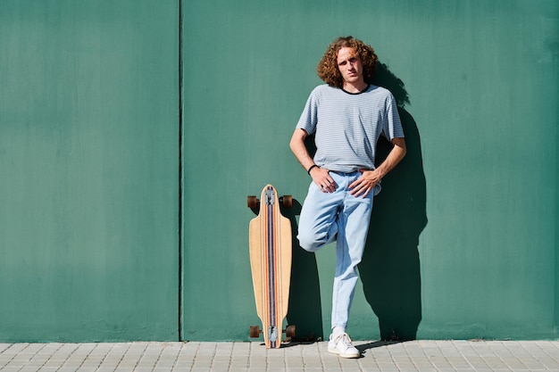 un giovane con i capelli ricci in piedi su un muro verde in una giornata di sole con il suo skate o longboard