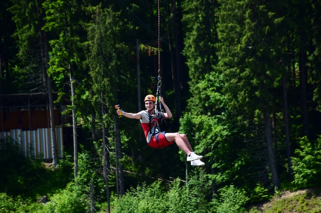 Un giovane con un casco è saltato dal bungee jumping e ora è appeso a una corda, oscillando e riprendendosi su una videocamera sportiva su uno sfondo sfocato di una foresta verde
