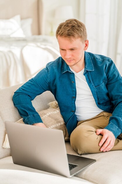 Un giovane con una camicia blu sta lavorando con un laptop mentre è seduto sul divano di casa