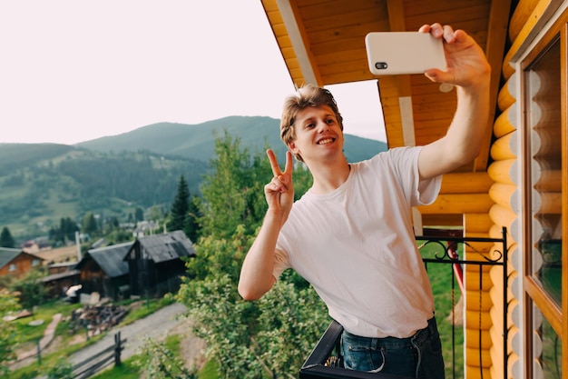 un giovane con una maglietta bianca e jeans si trova sul balcone di una casa collinare e si fa un selfie