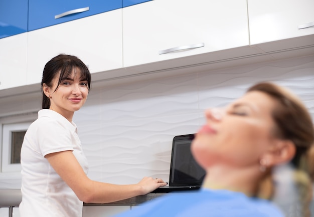 Un giovane dentista sorridente all'ufficio di odontoiatria che lavora al computer portatile e che consulta un paziente su una sedia dentale