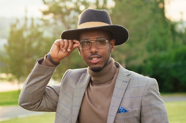 Un giovane e bell'uomo afroamericano modello alla moda in un elegante abito marrone e un cappello nero indossando occhiali in un parco estivo. latino americano ispanico uomo d'affari ragazzo nero foto modello.