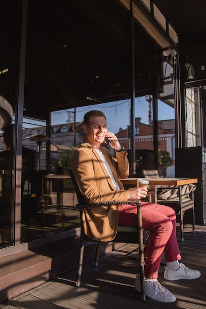 un giovane è seduto in un bar a bere caffè parlando al telefono e sorridendo