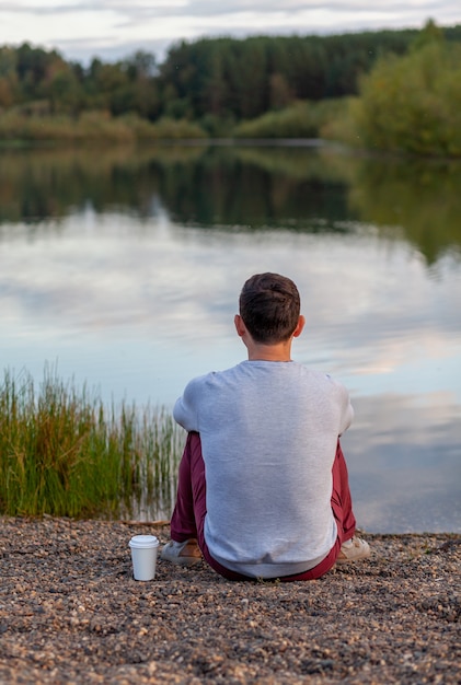 Un giovane è seduto sulla riva del lago e guarda lontano, accanto a una tazza bianca di caffè o tè. Solitudine e relax nella natura. Un bellissimo paesaggio di un lago nella foresta