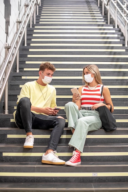 Un giovane e una donna che indossano maschere si siedono sulle scale di una stazione della metropolitana a parlare con i telefoni cellulari