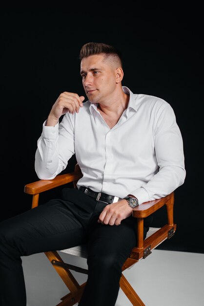 Un giovane elegante con una camicia bianca è seduto sulla sedia del regista su uno sfondo nero. Un bell'uomo d'affari carismatico.