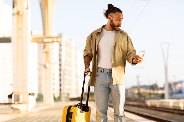 Un giovane europeo triste, confuso e scioccato, con la barba e la valigia, guarda l'orologio al telefono in ritardo