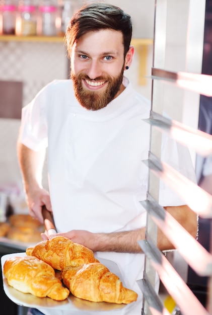 Un giovane fornaio maschio bello tiene un vassoio con cornetti francesi davanti a una panetteria e sorride.