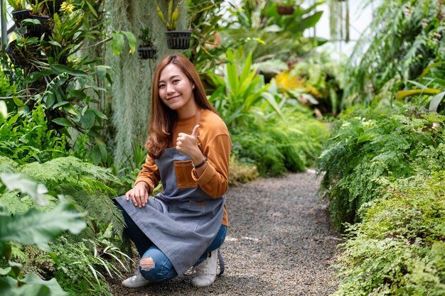 Un giovane giardiniere femminile asiatico che fa e mostra i pollici in su segno della mano mentre si prende cura delle piante nel giardino